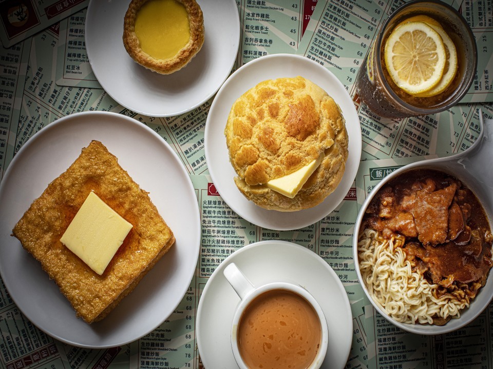 A table of cha chaan teng classics such as pineapple bun, egg tart, French toast, noodles, milk tea and lemon tea.