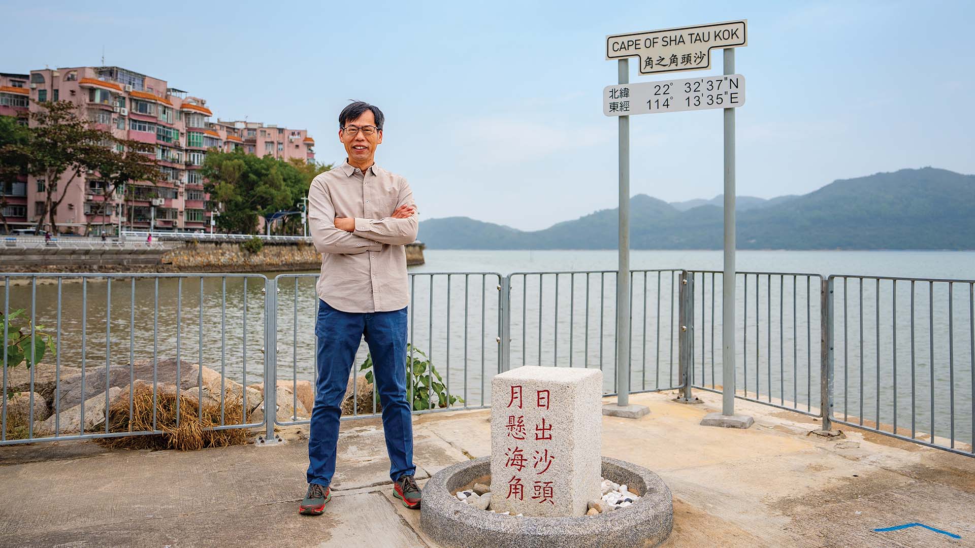 Charles Lee standing in front of the signpost “Cape of Sha Tau Kok”, surrounded by the panoramic coastal scenery.