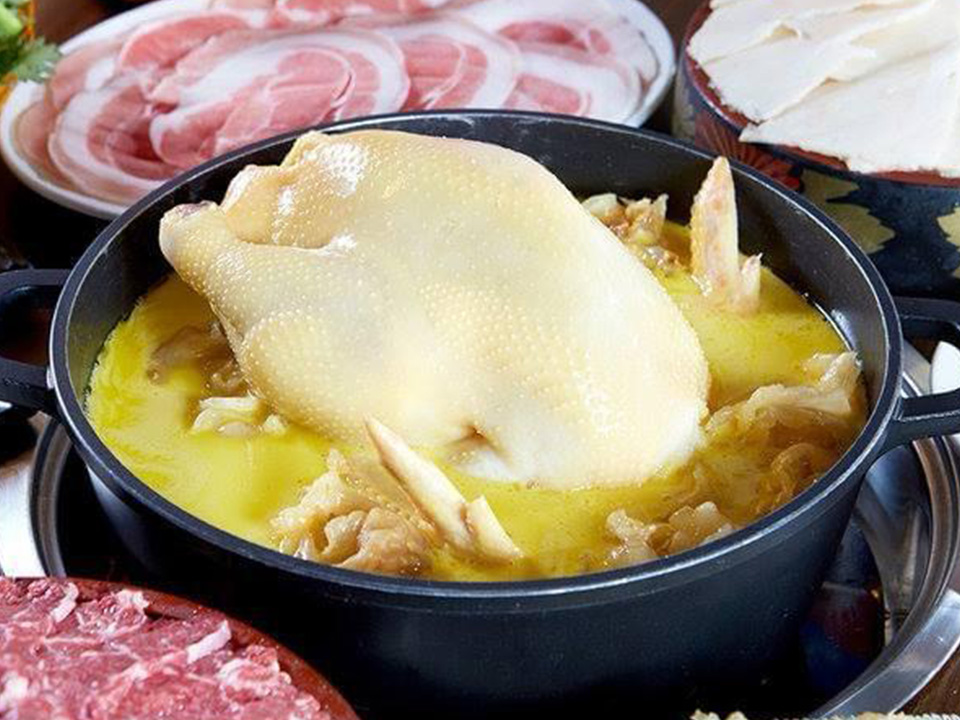 Large hotpot filled with a cooked chicken surrounded by plates of other meats